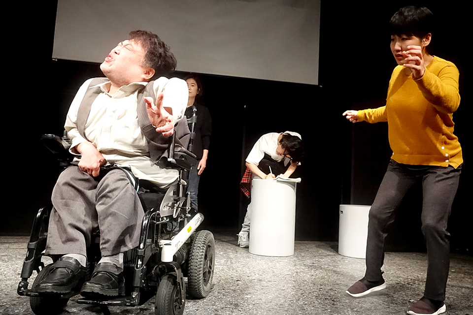 왼편에 휠체어를 탄 파우스트 역 배우가 괴이한 표정과 손동작을 하고 있고, 오른편에 그것을 바라보는 당황한 표정과 몸짓의 연출 역 배우, 뒤편으로 허리 높이의 작은 의자에 대고 대본에 뭔가를 적고 있는 조연출 역 배우가 보인다. 