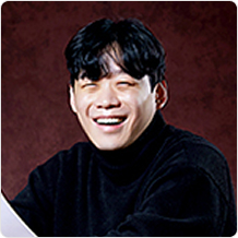 검은 스웨터를 입은 남성이 한 손에 종이를 들고 정면을 향해 환하게 웃고 있다.