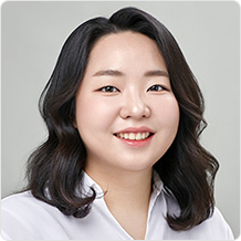 중단발 곱슬머리에 하얀색 셔츠를 입은 여성이 살짝 미소짓고 있다.