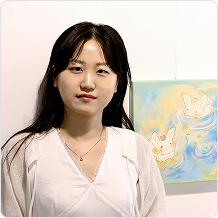 긴머리에 하얀색 원피스를 입은 여성이 우산토끼 그림 앞에 서서 정면을 향해 있다.