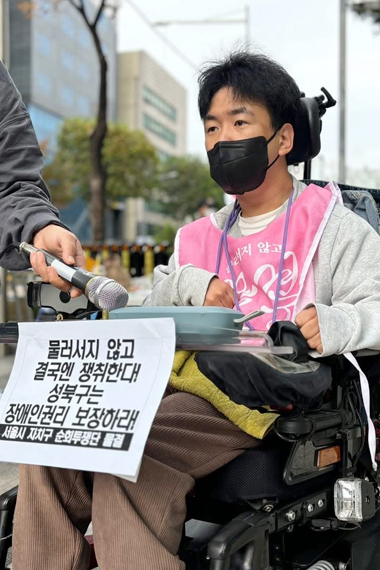 분홍색 피켓조끼를 입고 전동휠체어를 탄 홍성훈. 앞에는 “물러서지 않고 결국엔 쟁취한다!. 성북구는 장애인권리 보장하라! 서울 자치구 순회투쟁단 물결”이라고 쓰인 선전물이 부착되어 있다.