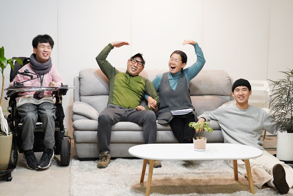깔끔한 흰색 벽면과 카페트가 깔린 공간에서, 회색 소파와 낮은 원형 탁자를 중심으로 네 사람이 휠체어에, 소파에, 바닥에 편하게 앉아 있다. 