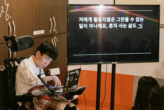 흰색 셔츠를 입은 홍성훈이 전동휠체어에 앉아 휠체어에 거치된 노트북 화면을 타이핑하고 있다. 오른쪽 큰 모니터에 “저에게 활동지원은 그만둘 수 있는 일이 아니에요, 혼자 사는 삶도 ㄱ”이라고 표시되고 있다.