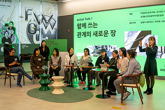 ‘Artist Talk 1 함께 쓰는 관계의 새로운 장’이라는 문구가 적힌 초록색 대형 스크린 무대 앞. 여러 명의 패널과 작가들이 원형으로 의자에 앉아 이야기를 나누고 있으며, 패널 뒤쪽 양 끝에서 수어통역사가 서서 수어를 하고 있다.