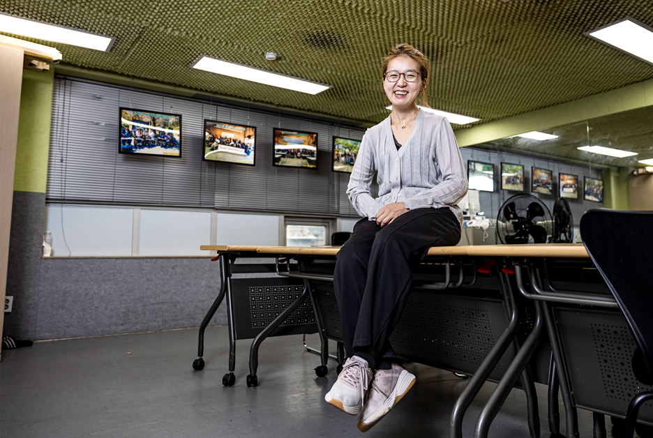안경을 쓴 박세영 배우가 책상 위에 앉아 전방을 바라보며 미소를 짓고 있다. 여성은 회색 가디건과 검은색 바지, 운동화를 착용하고 있다. 뒤편에는 블라인드가 내려져 있으며, 그 위로 여러 개의 사진 액자가 일정 간격으로 걸려 있다.