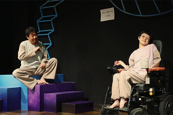 어두운 배경의 무대 위. 왼쪽에는 파란색과 보라색으로 칠해진 계단식 구조물 위에 남성 배우가 양반다리로 앉아 맞은편을 바라보고 있다. 오른쪽에는 안경을 쓴 김정희 배우가 전동 휠체어에 앉아 있다. 두 배우 모두 편안한 상하의 차림에 맨발 상태이다.